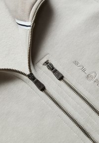 Ljusgrå zip-up jacka med två metallblixtlås och texturerad tyg. Inkluderar huva och broderad logotyp på framsidan.