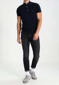 Granatowa koszulka polo z trzema guzikami, slim fit, w połączeniu z ciemnoniebieskimi dżinsami i szarymi sneakersami, dopełniona białymi skarpetkami.