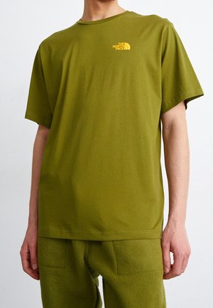 Osoba nosząca oliwkowo zielony t-shirt z krótkim rękawem i żółtym logo The North Face na piersi oraz pasujące teksturowane spodnie na jednolitym tle.
