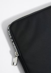 Funda negra para portátil con un exterior de tela suave, cremallera y herrajes plateados, que presenta un logo blanco en el borde de la cremallera.