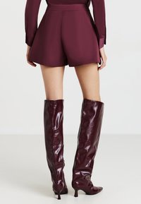 Donna che indossa pantaloni corti a vita alta color bordeaux e un top a maniche lunghe coordinato, abbinati a stivali con tacco lucidi alti fino al ginocchio color bordeaux, in piedi su un pavimento bianco.