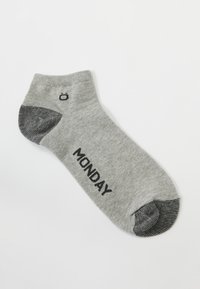 Graue Knöchelsocke mit schwarzen Akzenten an Zehen und Ferse. Schwarzer Text "MONTAG" auf der Seite gedruckt. Der Stoff hat eine glatte, gestrickte Textur.