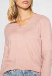 Helles, rosa V-Ausschnitt-Pullover mit einer strukturierten, fransigen Kantenhalslinie und langen Ärmeln, kombiniert mit blauen Jeans.