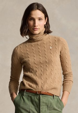 Rövid barna hajú nő, aki tevés színű, fonott mintás garbót, zöld nadrágot és fonott barna övet visel, kezét zsebében tartva.