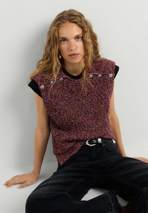 Jeune femme aux cheveux blonds ondulés portant un top rouge en maille sans manches avec des boutons et un jean noir, assise contre un mur clair uni.