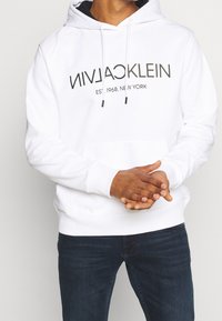 Valkoinen huppari, jossa on nyöri, musta logo ja teksti edessä, pehmeä materiaali ja kengurutasku. Yhdistetty tummiin farkkuihin.