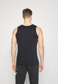Man staat met zijn rug naar ons toe, gekleed in een zwart mouwloos tanktop en zwarte broek met witte strepen aan de linkerzijde.