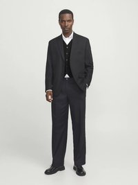 Černé sako přes černou vestičku, bílá košile a tmavé kalhoty. Texturovaná látka s přizpůsobeným střihem, doplněná elegantními botami.