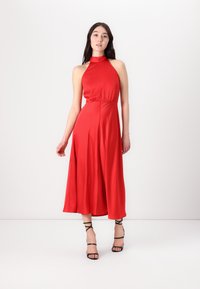 Robe rouge à col halter avec un corsage ajusté, une jupe mi-longue fluide et une fente sur le côté. Associée à des sandales à talons hauts noires à lanières.