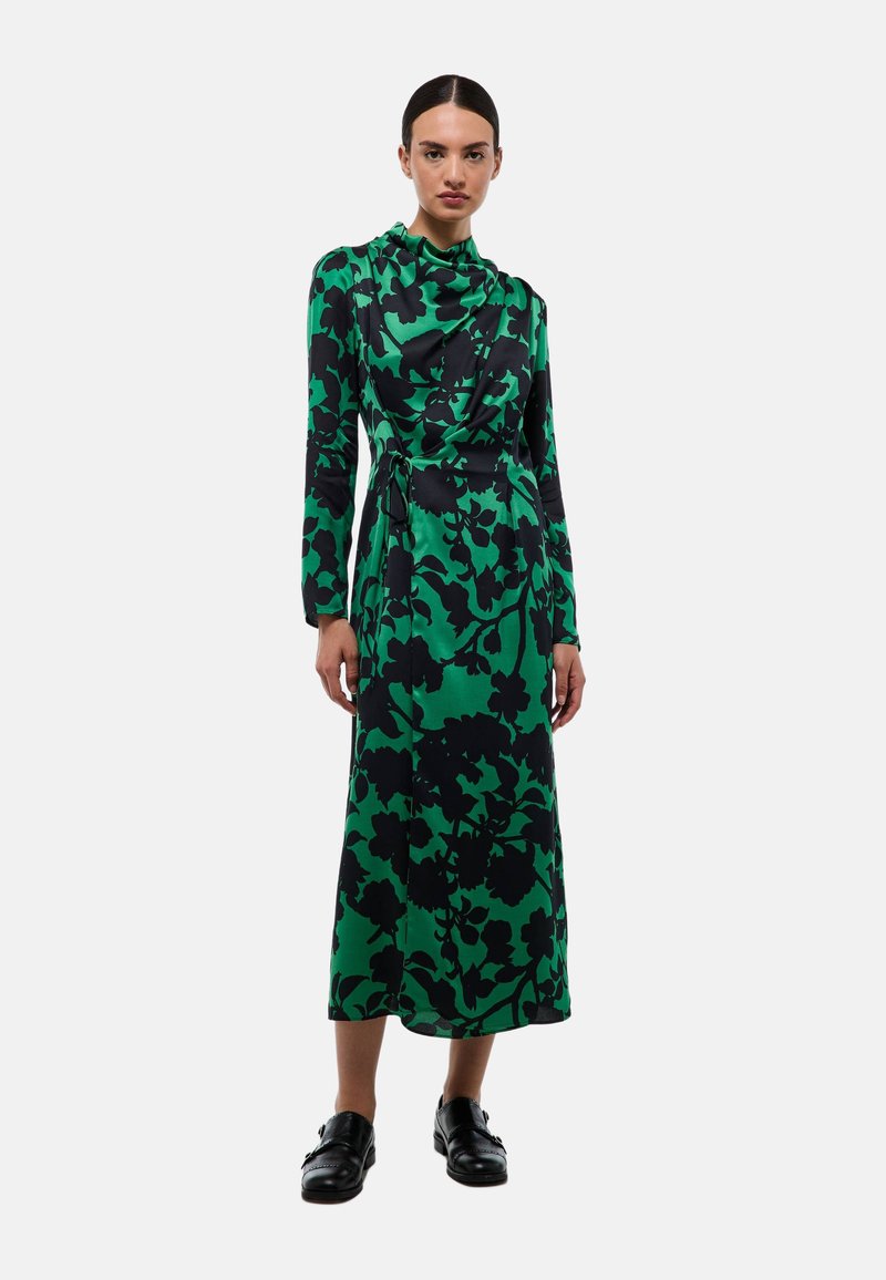 Robe verte à manches longues avec un motif floral noir, design wrap et encolure froncée. Portée avec des chaussures noires, elle présente une coupe mi-longue.