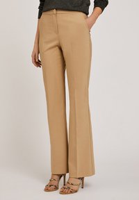 Pantalons évasés beiges en tissu lisse, dotés d'une fermeture éclair à l'avant, de passants de ceinture et de poches latérales. Portés avec des sandales à brides.