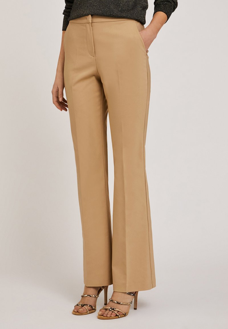 Pantalons évasés beiges en tissu lisse, dotés d'une fermeture éclair à l'avant, de passants de ceinture et de poches latérales. Portés avec des sandales à brides.