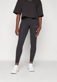 Černé legíny s vysokým pasem vyrobené z elastického materiálu, přiléhavý střih a kontrastní bílé logo na levém stehně. Spárovány s oversized černým topem.