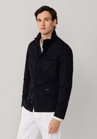 Hombre con chaqueta negra con cremallera y bolsillos de solapa, y pantalones blancos, de pie contra un fondo liso de color gris claro.