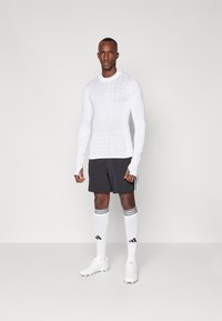 Weißes langärmliges Sportoberteil mit Karomuster, kombiniert mit anthrazitfarbenden Shorts, weiß-schwarz gestreiften Socken und weißen Fußballschuhen.