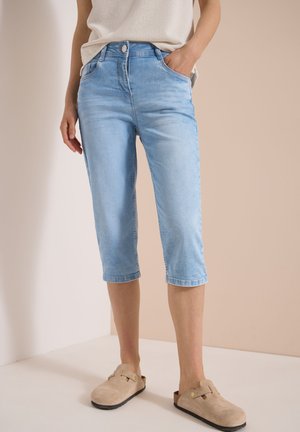Kvinde iført lyseblå denim capri bukser, beige slip-on sko og en cremefarvet strikket top, stående mod en beige og hvid baggrund.