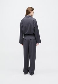 Γυναίκα με μακριά καστανά μαλλιά, γυρισμένη πλάτη, φοράει χαλαρό σκούρο γκρι σακάκι και αντίστοιχο παντελόνι με φαρδιά μπατζάκια, σε λευκό απλό φόντο.