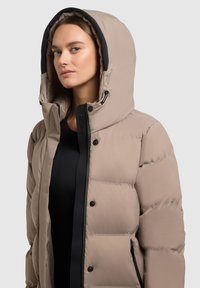 Puffer jacket in light beige with a black interior, featuring a large hood, snap buttons, and quilted texture. Zippered pocket visible.