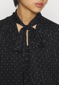 Blusa nera leggera con dettaglio al collo a fiocco, caratterizzata da righe verticali e piccoli punti bianchi. Apertura a chiave sul colletto.