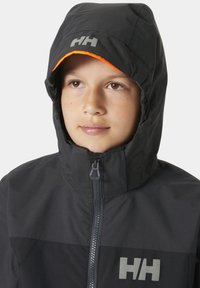 Czarna kurtka z kapturem z szarym logo, z zamkiem błyskawicznym z przodu i pomarańczowymi wykończeniami na kapturze. Materiał wydaje się gładki i odporny na warunki atmosferyczne.