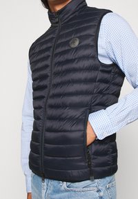 Gilet trapuntato navy con colletto alto, chiusura frontale con zip e tasche laterali. Abbinato a una camicia a quadri azzurri chiari sotto.