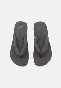 Svarta flip-flops med vadderade tåringar och texturerad sula, med en diskret logotypprägling på fotbädden. Enkel, minimalistisk design.