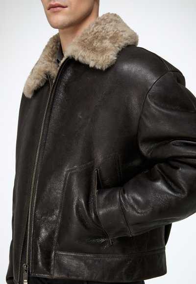 Man wearing a dark brown leather jacket with a beige shearling collar, hand in zippered pocket, against a plain white background.