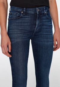 Dark blue denim jeans with a slim fit, featuring a classic five-pocket design and visible stitching details. Smooth texture and button closure.