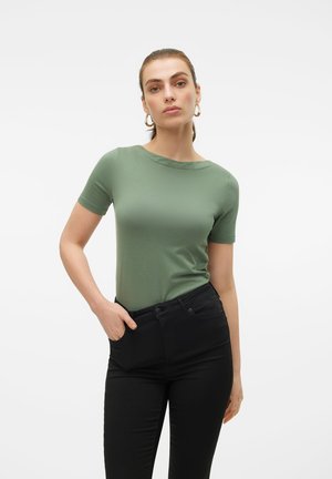 Femme portant un haut vert ajusté à manches courtes et un pantalon noir taille haute, debout avec une main dans la poche sur fond blanc.