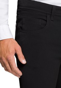 Black stretch jeans with a slim fit, featuring a small front pocket, detailed stitching, and a mid-rise waistband. Hand resting on the fabric.