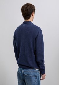 Man facing away wearing a dark blue sweater and light blue jeans against a plain light gray background.