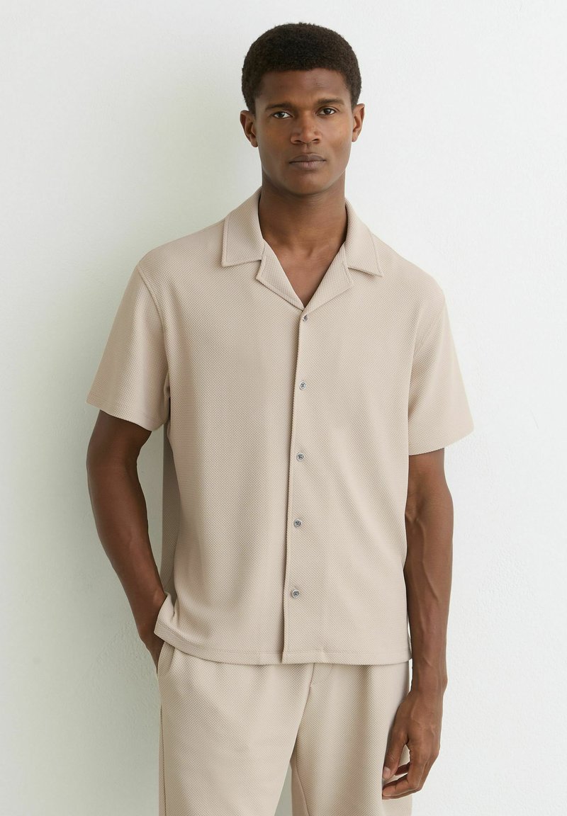 Hombre con camisa beige de manga corta con botones y pantalones a juego, de pie contra una pared blanca lisa, con las manos relajadas.