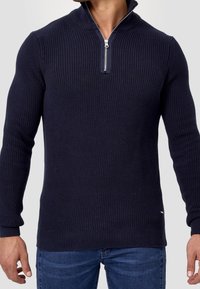 Granatowy sweter z prążkowanego materiału z ćwierć zamkiem przy kołnierzu, długimi rękawami i teksturowanym splotem. Posiada mały detal z logo na dole.