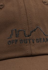 Brun stofkasket med sort broderet bysilhuet og teksten "OFF DUTY GEAR" på frontpanelet.