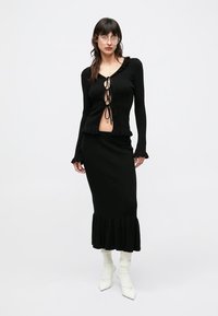 Juoda, raištuota megzta dviejų dalių apranga, kurios viršus yra trumpas ir su raukiniais, o ilgas sijonas turi raukinių apvadą. Derinama su baltais batais.