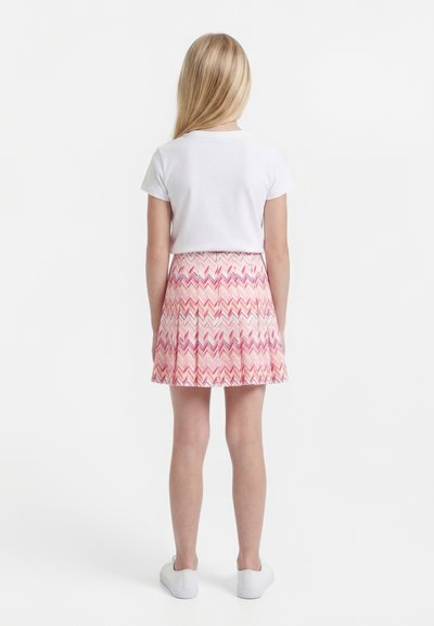 Fată cu păr blond lung, cu fața spre spate, purtând o cămașă albă cu mâneci scurte, fustă roz cu model și pantofi sport albi pe un fundal alb.