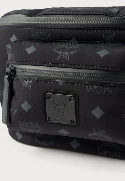 Mala preta texturizada com logótipo MCM, vários compartimentos com fecho éclair e padrões em relevo. Apresenta uma placa de metal preta com o logótipo e detalhes.