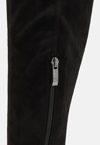 Black suede knee-high boot with a silver zipper detail on the side and visible stitching, showcasing a textured, soft surface.