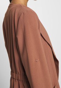 Dammig rosröd jacka med en texturerad tyg, utrustad med en dragsko i midjan och en knappdetalj på ärmen.