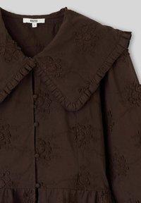 Blouse marron foncé avec un grand col à volants, des boutons recouverts de tissu et des motifs floraux brodés sur toute la surface.