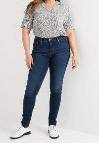 Mujer vestida con una blusa de manga corta estampada, jeans ajustados de color azul oscuro y zapatos blancos con cordones, de pie contra un fondo liso.