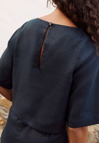 Dark navy top featuring a boxy design, short sleeves, and a keyhole opening at the back secured with a button. Smooth, textured fabric.