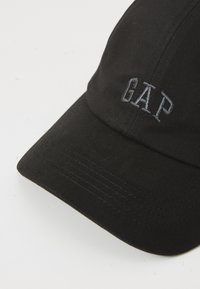 Casquette de baseball noire à visière incurvée avec un logo "GAP" brodé en gris sur le panneau avant.