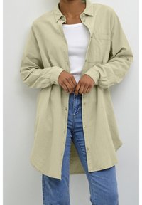 Chemise vert clair boutonnée avec col, manches longues et une poche poitrine, portée par-dessus un débardeur blanc et un jean bleu.