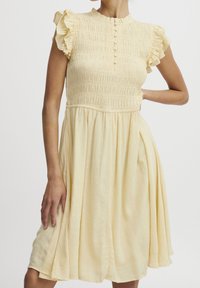 Helles gelbes Smock-Dress mit kurzen Volantärmeln, strukturiertem Stoff, tailliertem Oberteil und weit ausgestelltem Rock. Mit Knopfdetail an der Vorderseite.