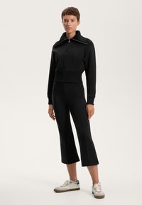 Mustahi jakkua musta, jolla on korkea kaulus, yhdistettynä mustiin tiukkoihin housuihin. Kengissä on vaalea pohja ja yksityiskohtia.