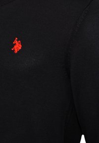 Plano detalle de una tela de punto negra con un pequeño logo bordado de un jugador de polo en rojo en el área del pecho.