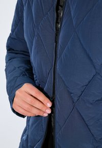 Mörkblå quiltad jacka med dragkedja fram; har en slät yta och diamantmönster; handen syns justera dragkedjan.