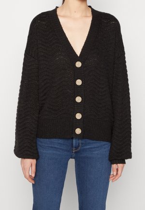 Žena oblečená v černém pleteném kardiganu s velkými béžovými knoflíky a modrých džínách, zobrazena od krku po boky, na bílém pozadí.