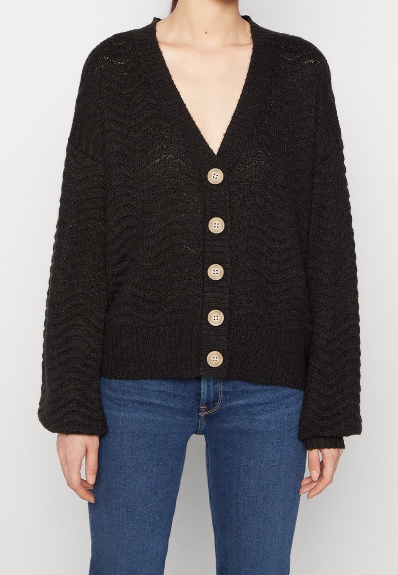 Vrouw draagt een zwarte gebreide cardigan met grote beige knopen en een blauwe spijkerbroek, gefotografeerd van nek tot heupen tegen een witte achtergrond.
